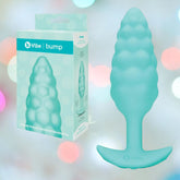 The B-Vibe Texture Vibrating Butt Plug Bump (Small, Mint) sits by its box. Made from body-safe silicone with a flared base for comfort and safety, it’s perfect for playful anal fun. Pastel bokeh lights add a whimsical touch to the scene.